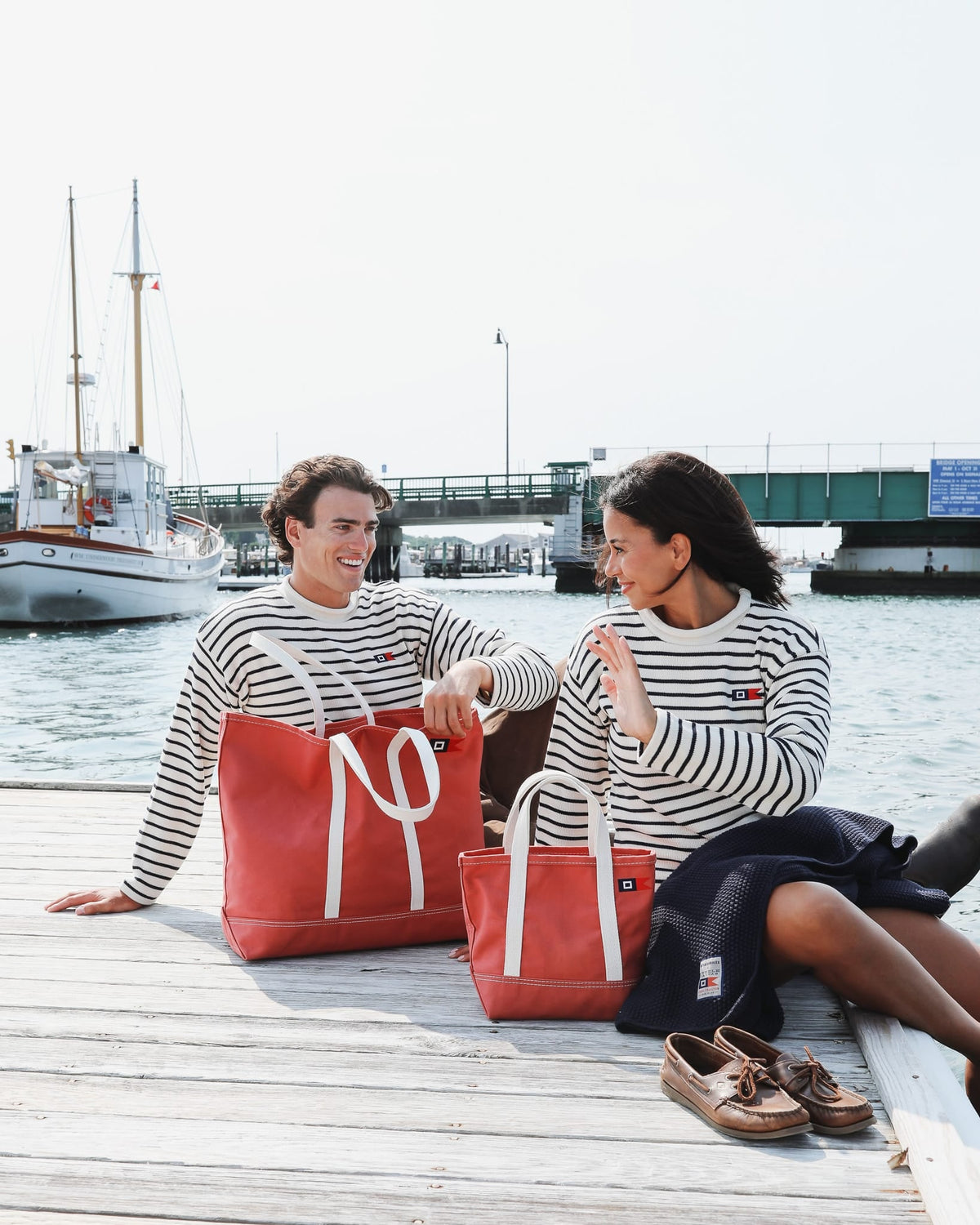 Man and woman in Signal Rollneck Sweaters sitting on a dock with Signal totes and the navy Signal Throw