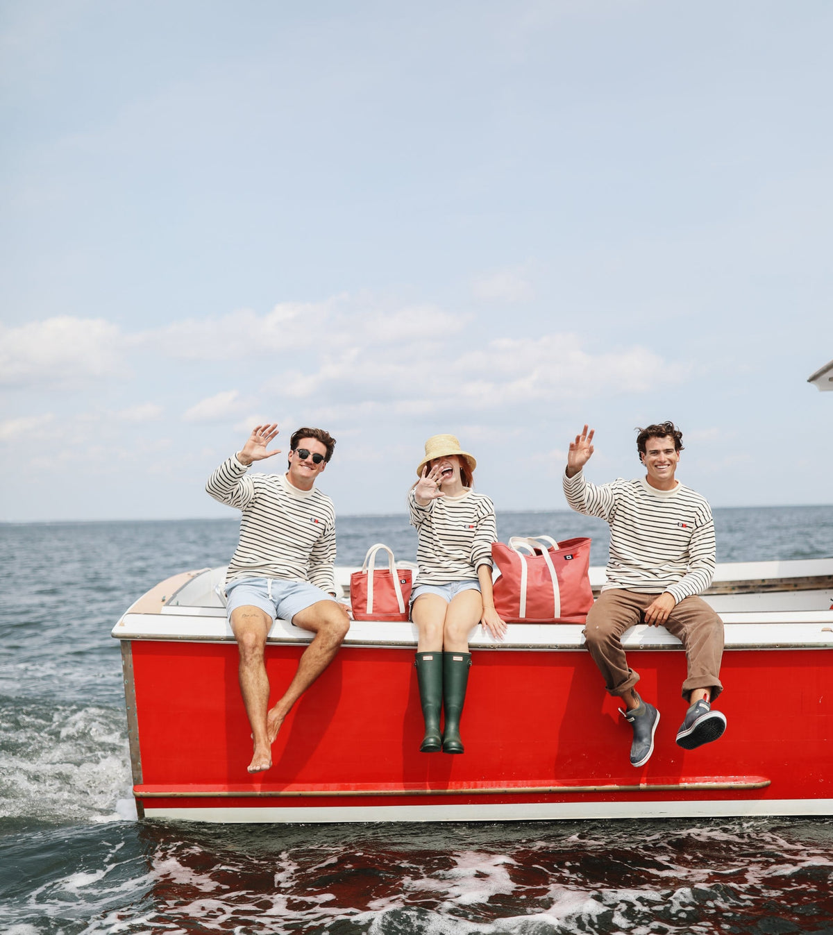 Three people in Signal Rollneck Sweaters wave from the side of the red lobster boat, with Signal totes between them