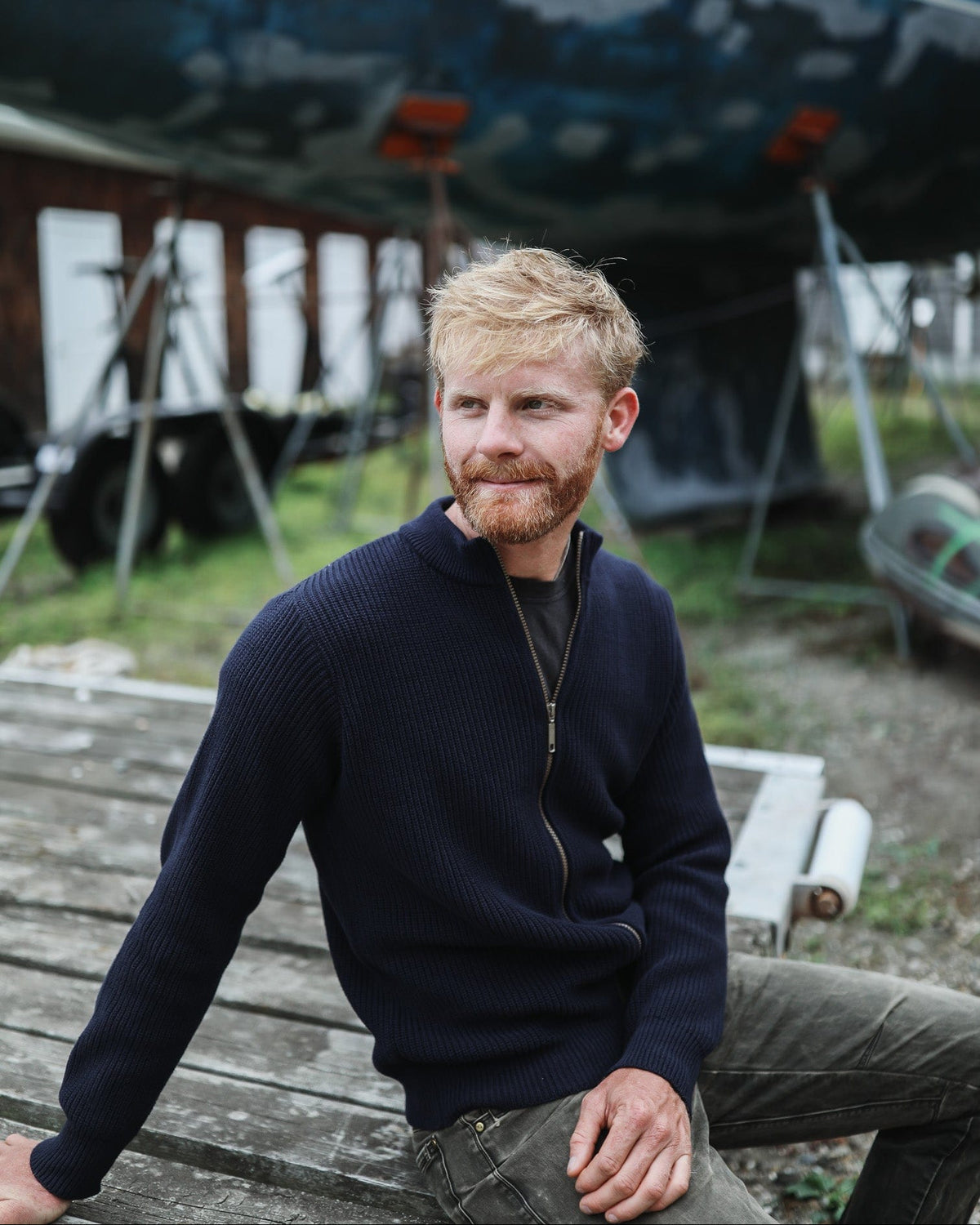 Whalerknits Cuttyhunk Full-Zip Cardigan: A man in navy and dark trousers sits on a boatyard bench.