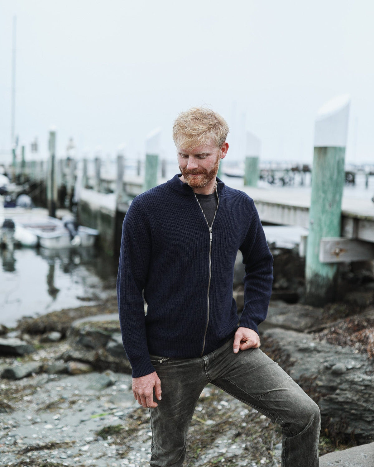 Whalerknits Cuttyhunk Full-Zip Cardigan: A man in navy stands on a rocky shore beside a dock.