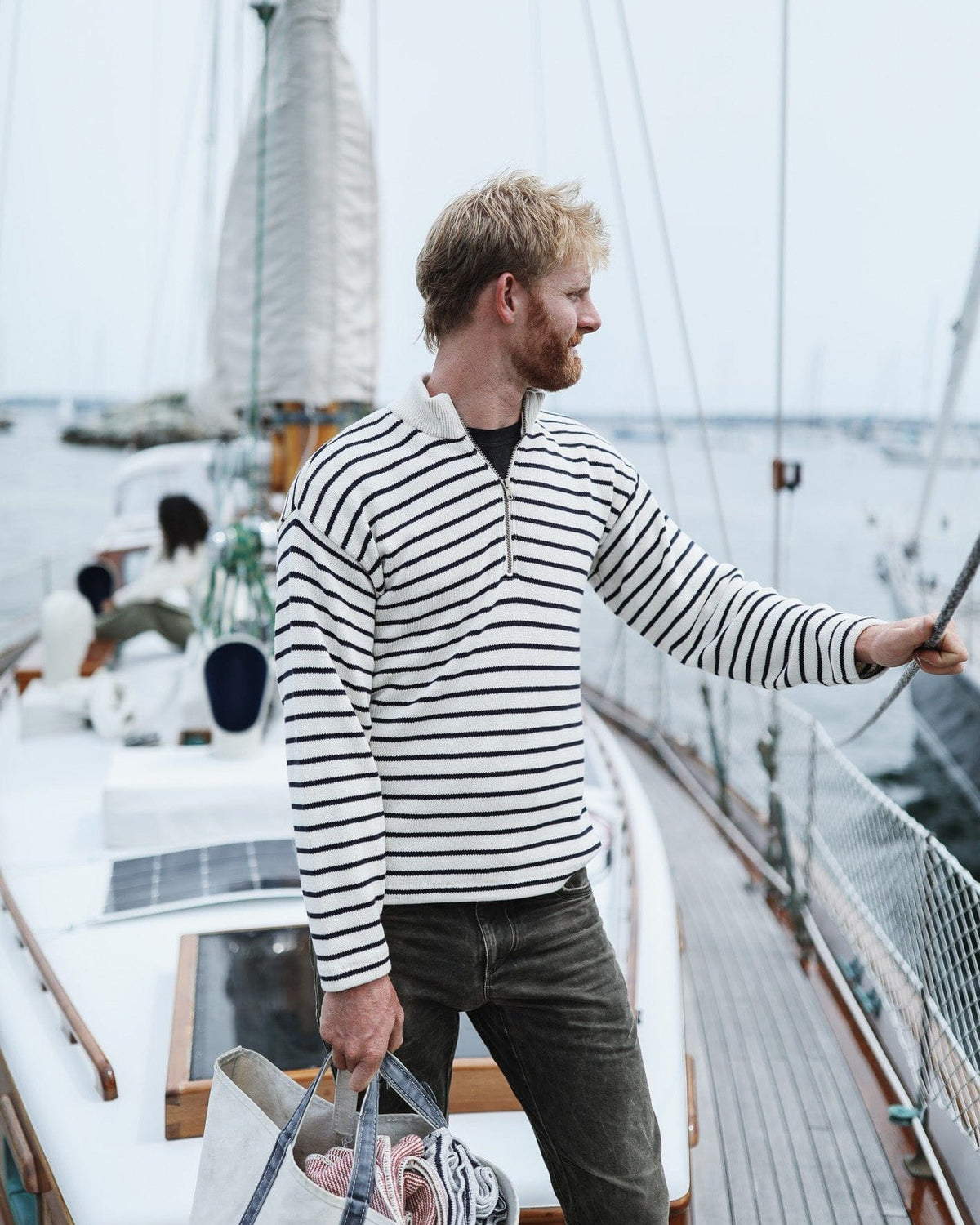 Whalerknits Nantucket Classic 1/4 Zip Sweater: A man in natural with navy stripes stands aboard a sailboat holding rigging and a patterned bag.