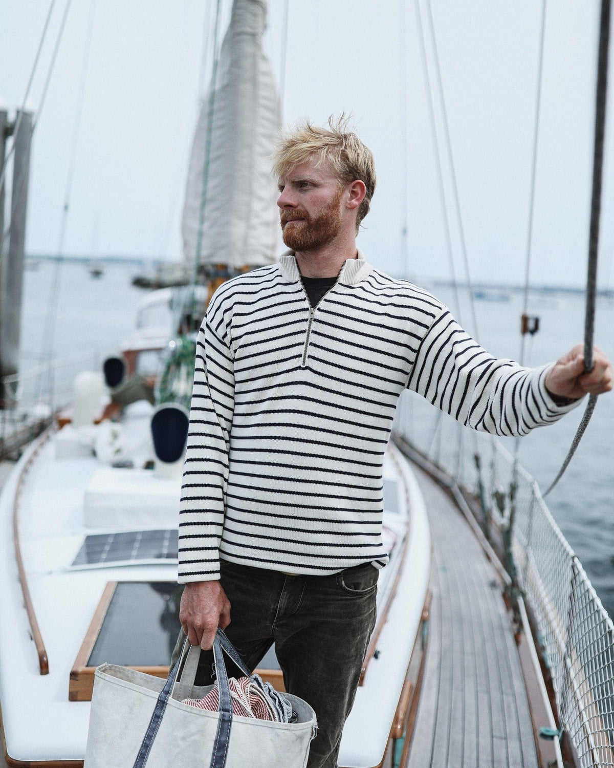 Whalerknits Nantucket Classic 1/4 Zip Sweater: A man in natural with navy stripes holds sailboat rigging and carries a patterned bag.