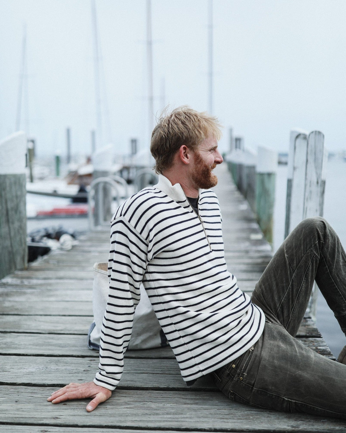 Whalerknits Nantucket Classic 1/4 Zip Sweater: Side view of a man in natural with navy stripes seated on a wooden dock.