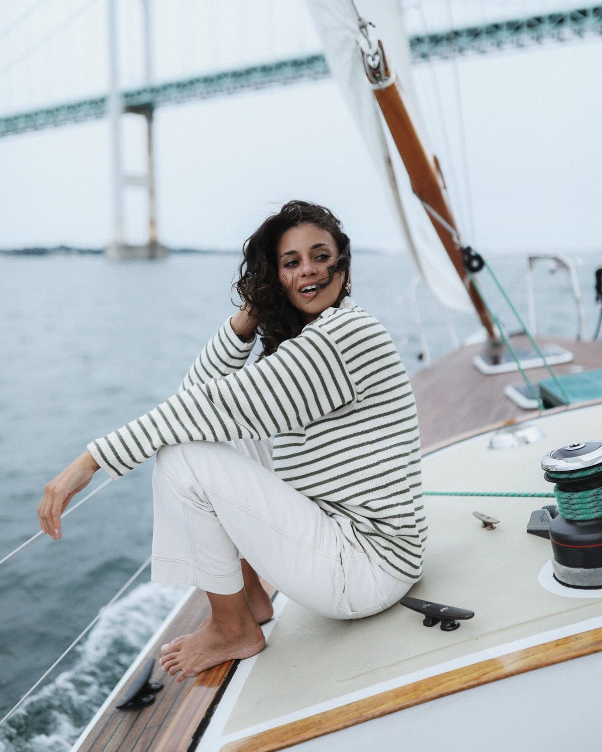 Whalerknits Nantucket Classic 1/4 Zip Sweater: A woman in natural with green stripes and white trousers sits barefoot beside a sailboat winch.