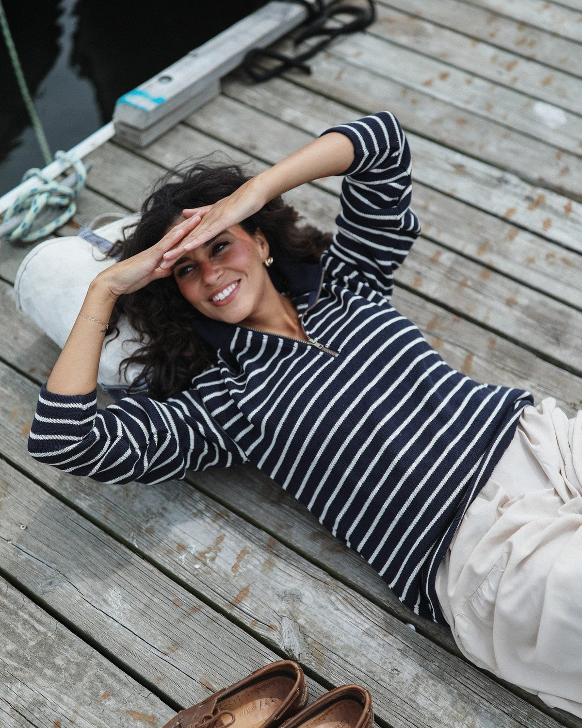 Whalerknits Nantucket Classic 1/4 Zip Sweater: A woman in navy with natural stripes reclines on a wooden dock with her arms raised.