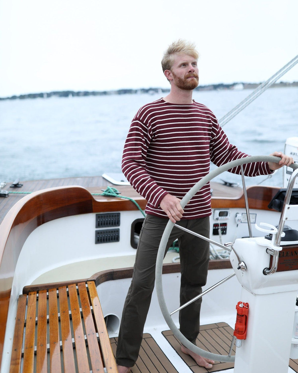 Whalerknits Nantucket Classic Boatneck Sweater: A man in burgundy with natural stripes stands at a sailboat wheel.