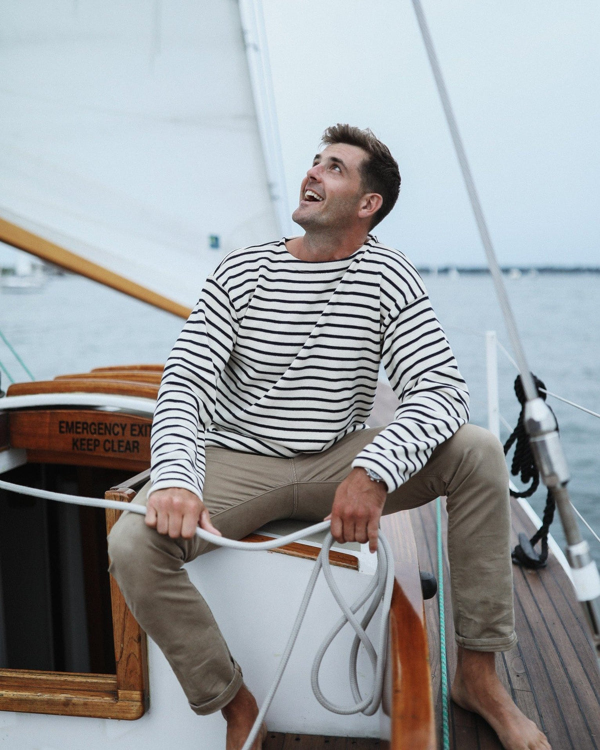 Whalerknits Nantucket Classic Boatneck Sweater: A man in natural with navy stripes sits holding a rope aboard a sailboat.