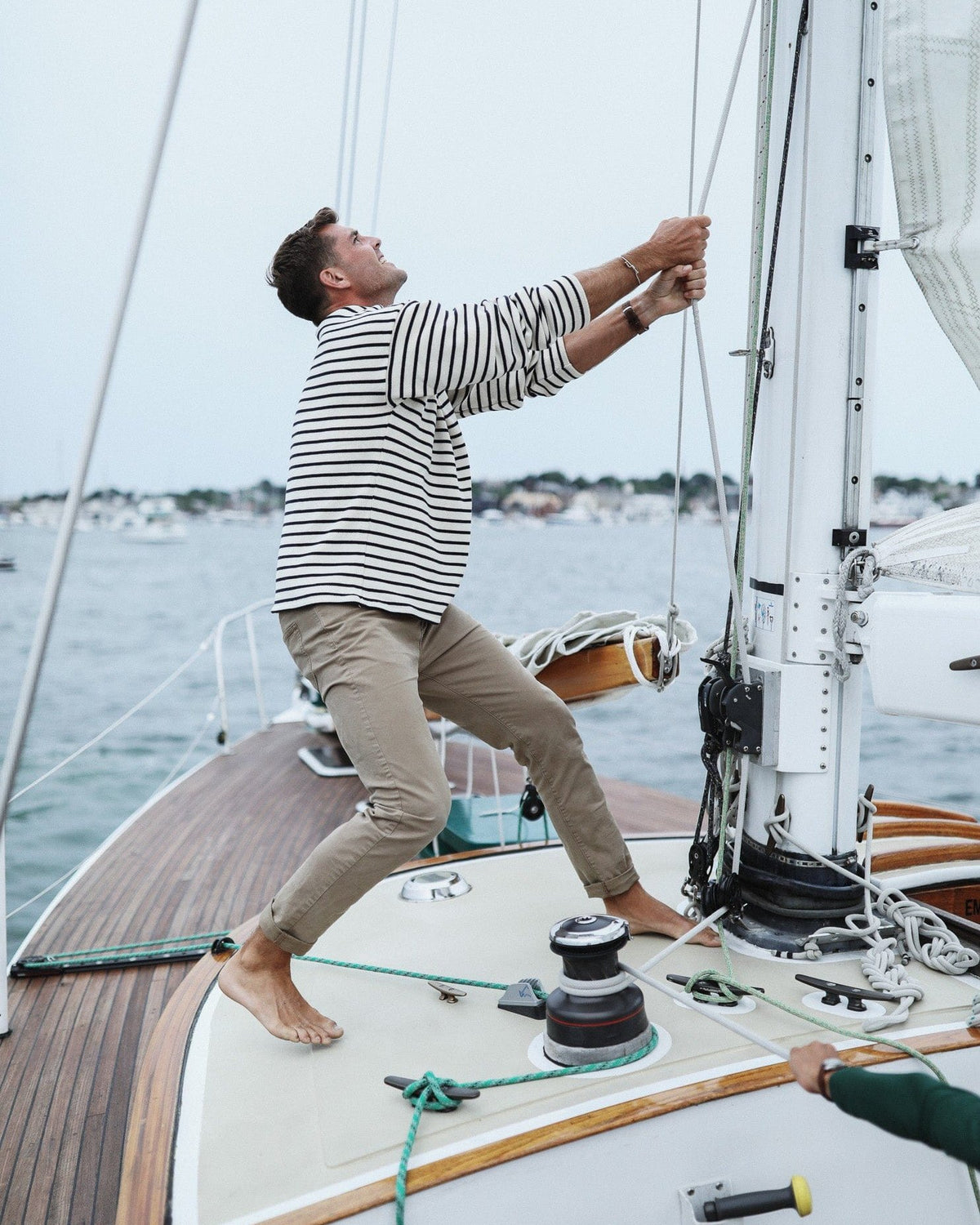 Whalerknits Nantucket Classic Boatneck Sweater: A man in natural with navy stripes pulls rigging while standing barefoot on a sailboat.