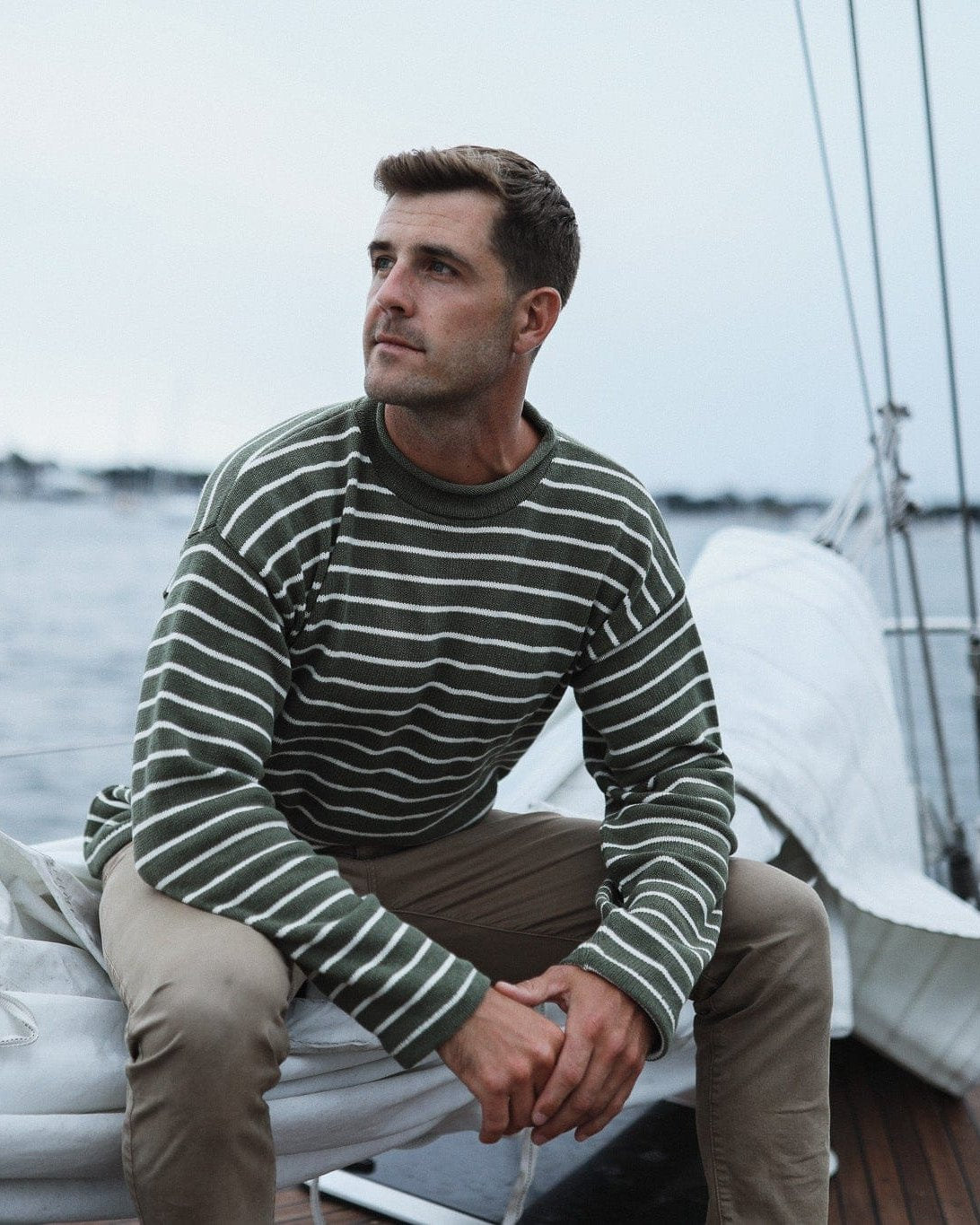 Whalerknits Nantucket Rollneck Sweater: A man in olive with natural stripes sits on folded sailcloth aboard a boat.