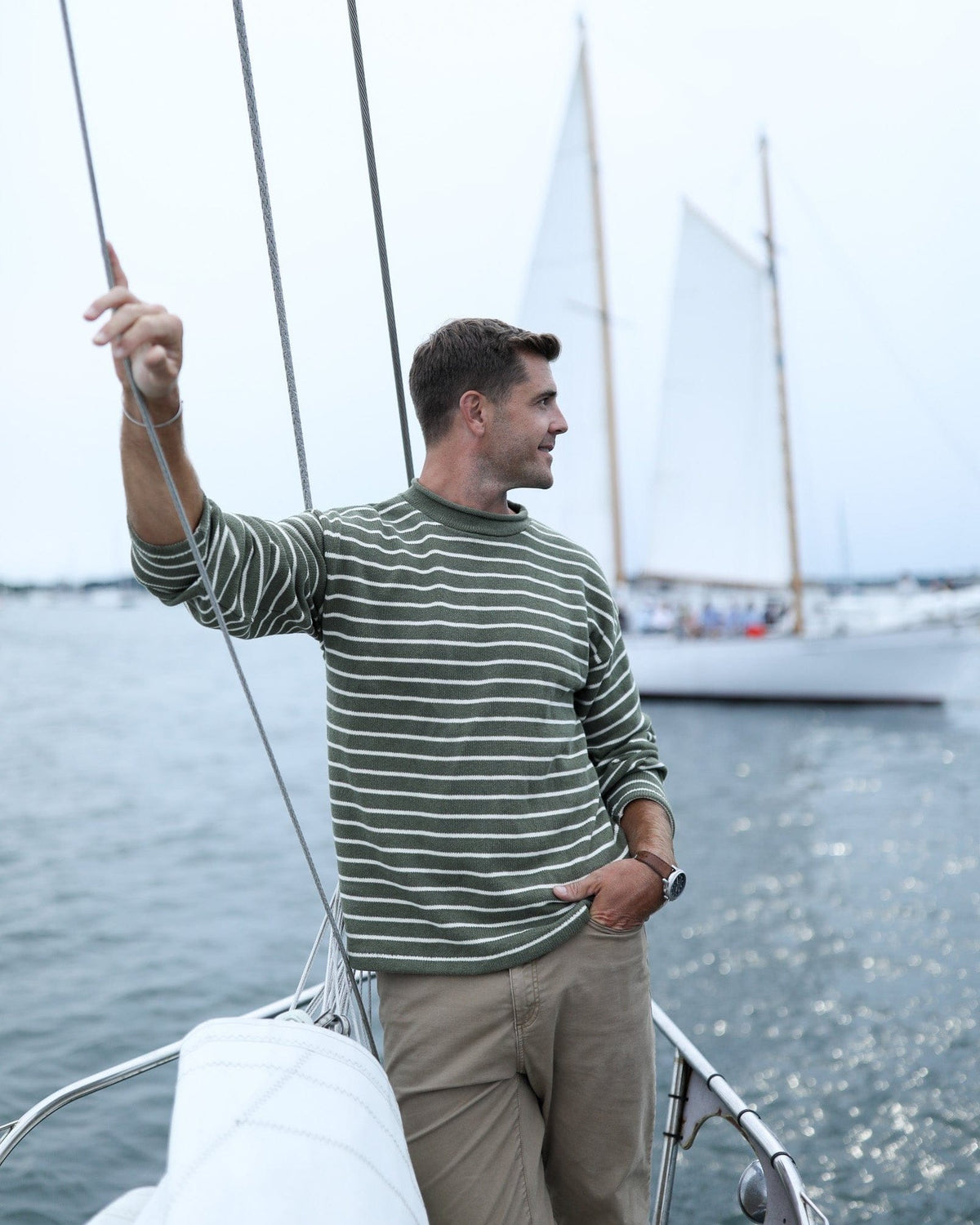 Whalerknits Nantucket Rollneck Sweater: A man in olive with natural stripes holds sailboat rigging, wearing tan trousers.