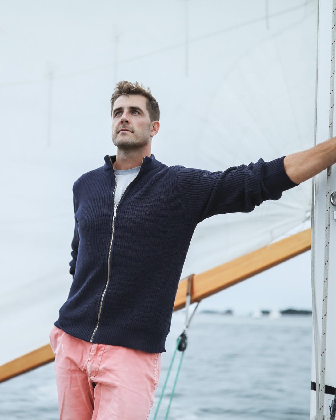 Whalerknits Cuttyhunk Full-Zip Cardigan: A man in navy and pink shorts holds rigging aboard a sailboat.