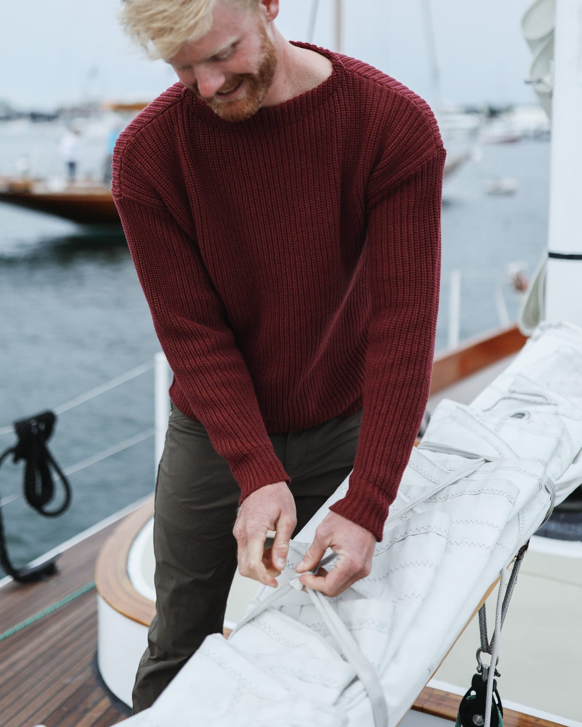 Whalerknits Newport Shaker Boatneck Sweater: A man in burgundy bends over sailcloth while working on a sailboat.