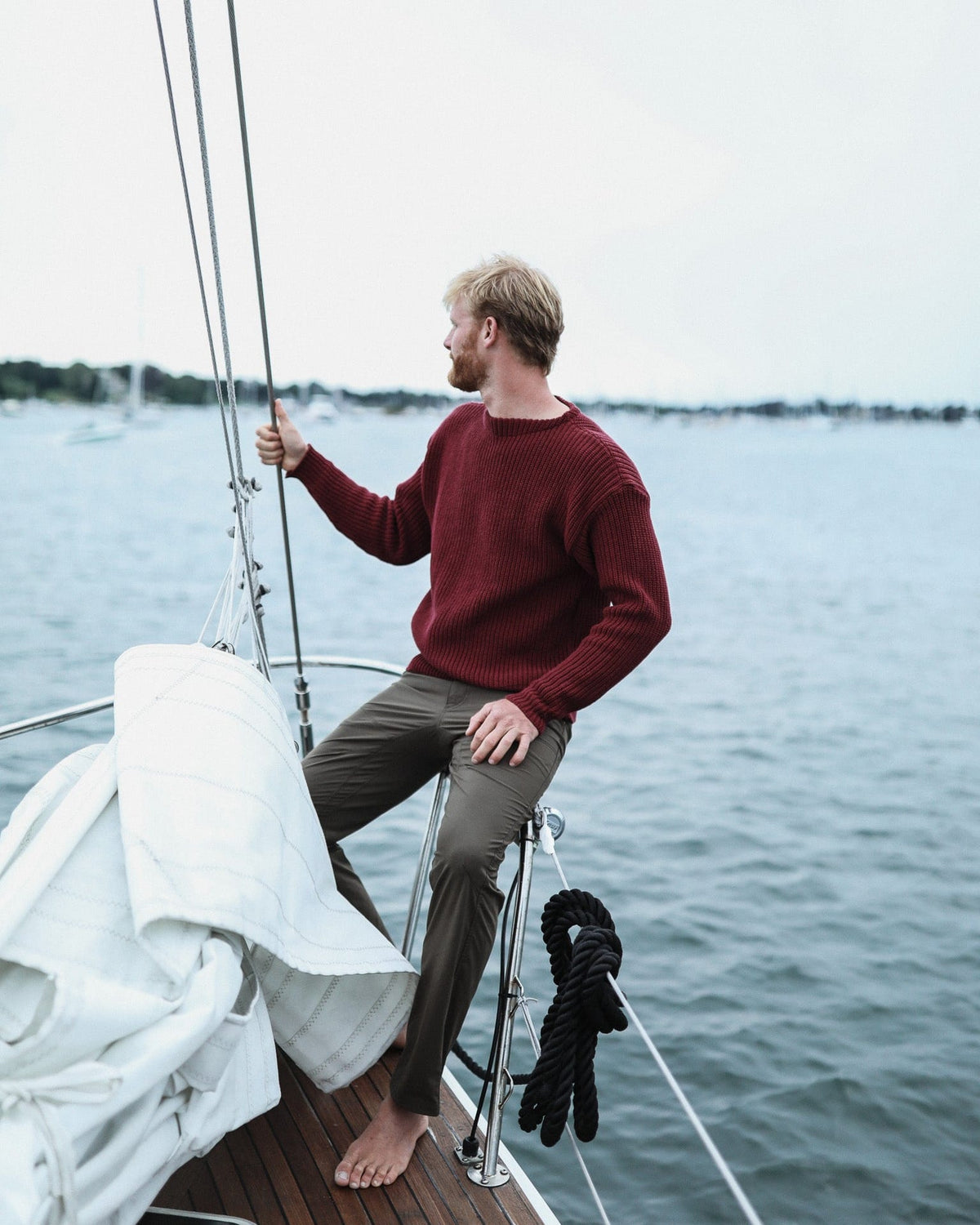 Whalerknits Newport Shaker Boatneck Sweater: A man in burgundy and dark trousers sits on a sailboat holding the rigging.