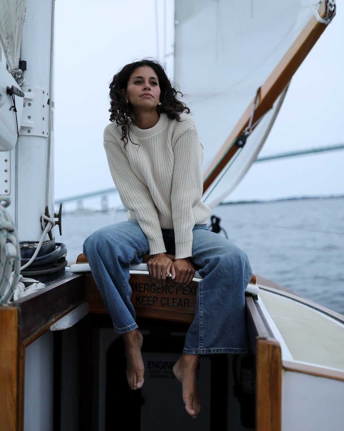 Whalerknits Newport Shaker Knit Crewneck Sweater: A woman in natural and jeans sits barefoot beside a sailboat mast.