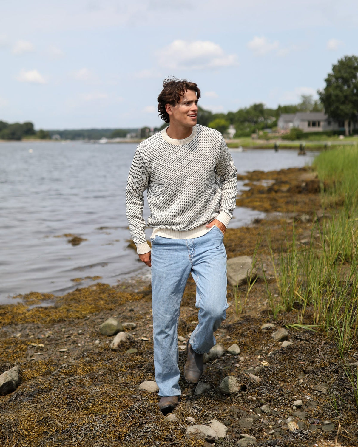 Whalerknits Sakonnet Birdseye Crewneck Pullover: A man in natural with navy birdseye pattern and jeans walks along a rocky shoreline.