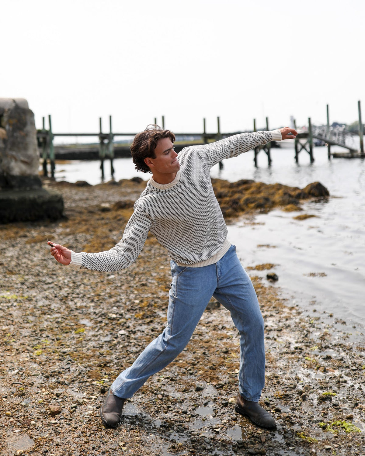 Whalerknits Sakonnet Birdseye Crewneck Pullover: A man in natural with navy birdseye pattern extends his arms on a rocky shoreline.
