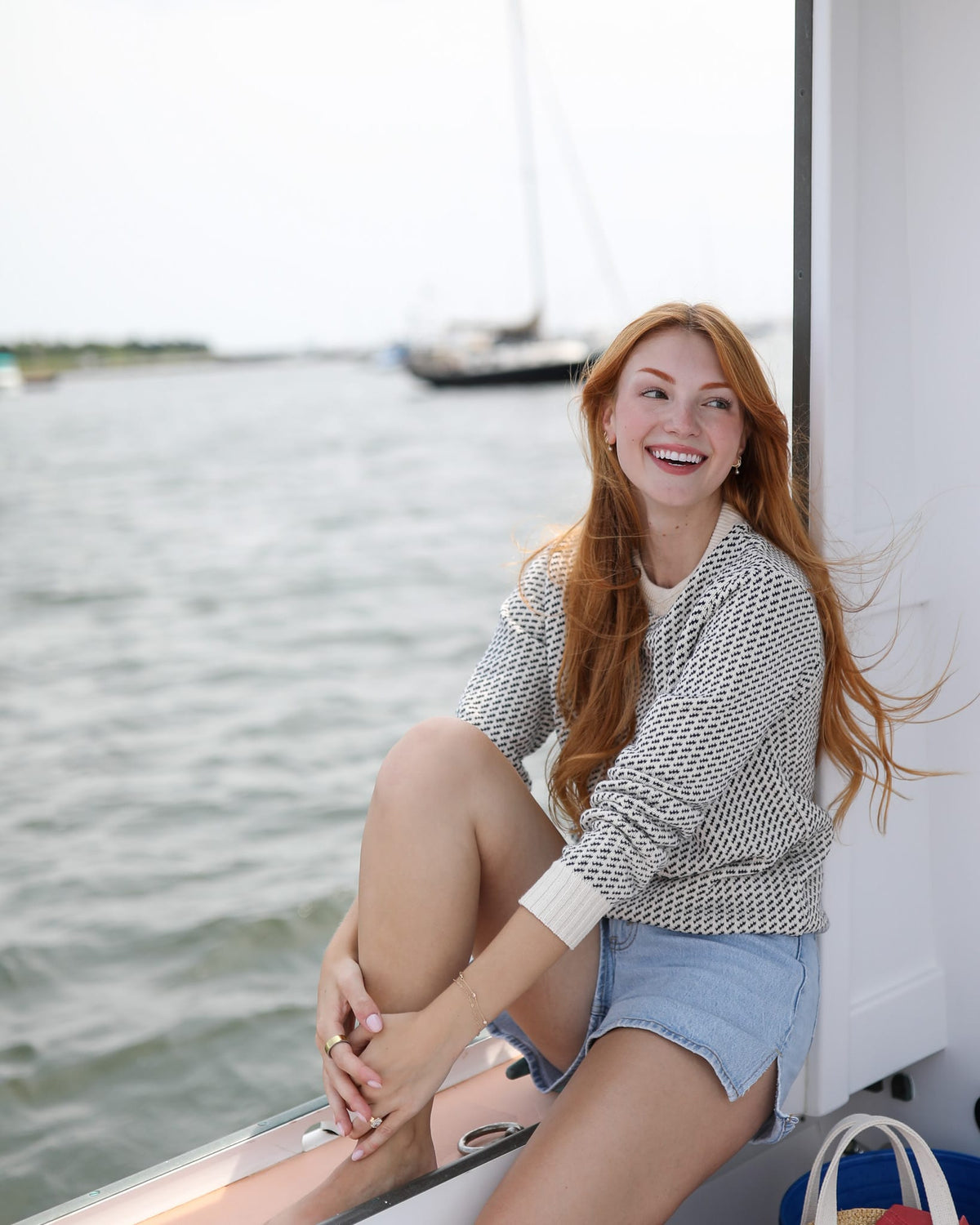 Whalerknits Sakonnet Birdseye Crewneck Pullover: A woman in natural with navy birdseye pattern and denim shorts sits smiling on a boat rail.