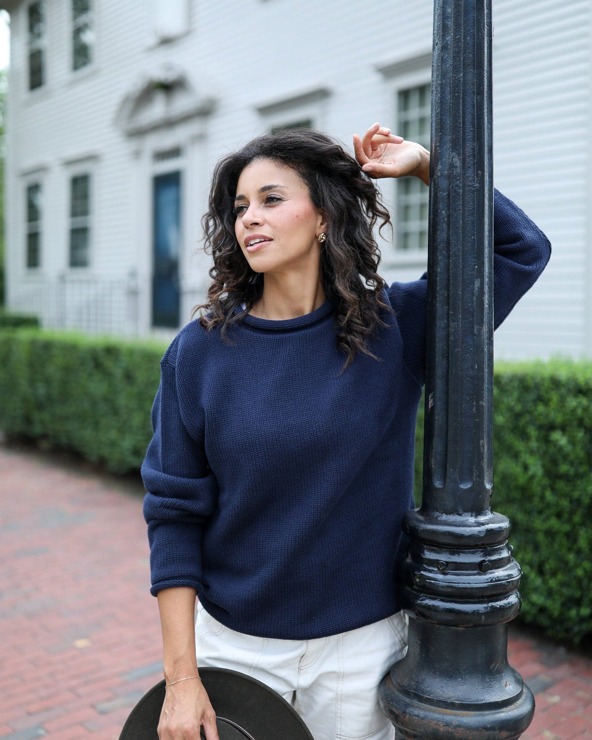 Whalerknits Wellfleet Rollneck Sweater: A woman in navy and white trousers leans against a lamppost on a brick sidewalk.
