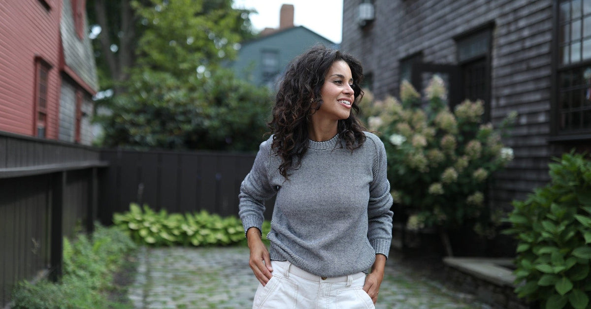 Whalerknits Westport Crewneck Saddle Shoulder Pullover: A woman in the gray fine-striped sweater and white trousers stands on a cobbled path between houses.