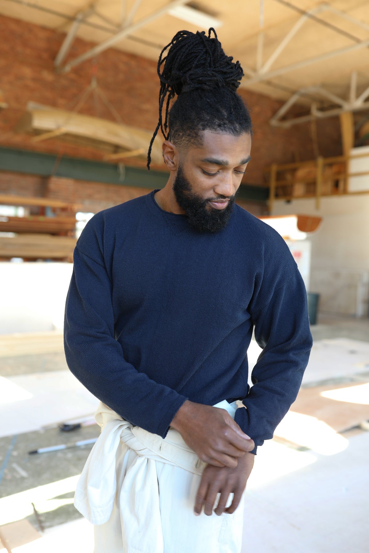 Whalerknits Chatham Jersey Sweatshirt Pullover: A man in navy adjusts his sleeve inside a boat workshop, with a white garment tied around his waist.