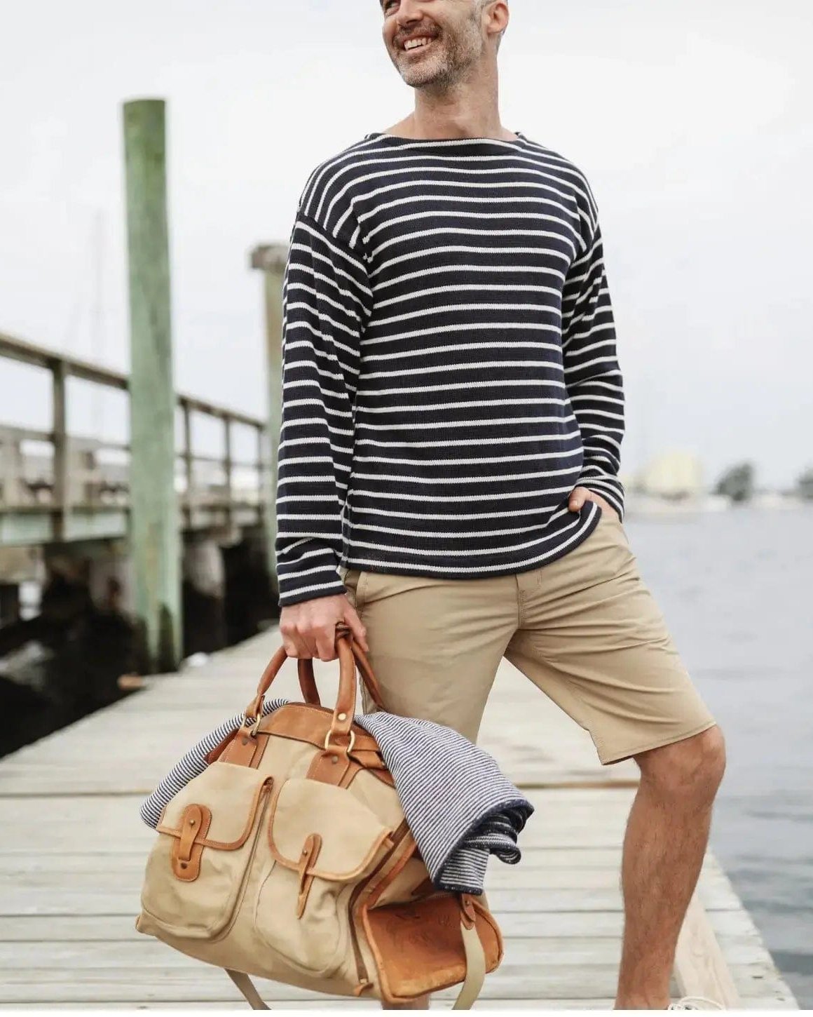 Whalerknits Nantucket Classic Boatneck Sweater: A man in navy with natural stripes and tan shorts carries a canvas bag on a dock.