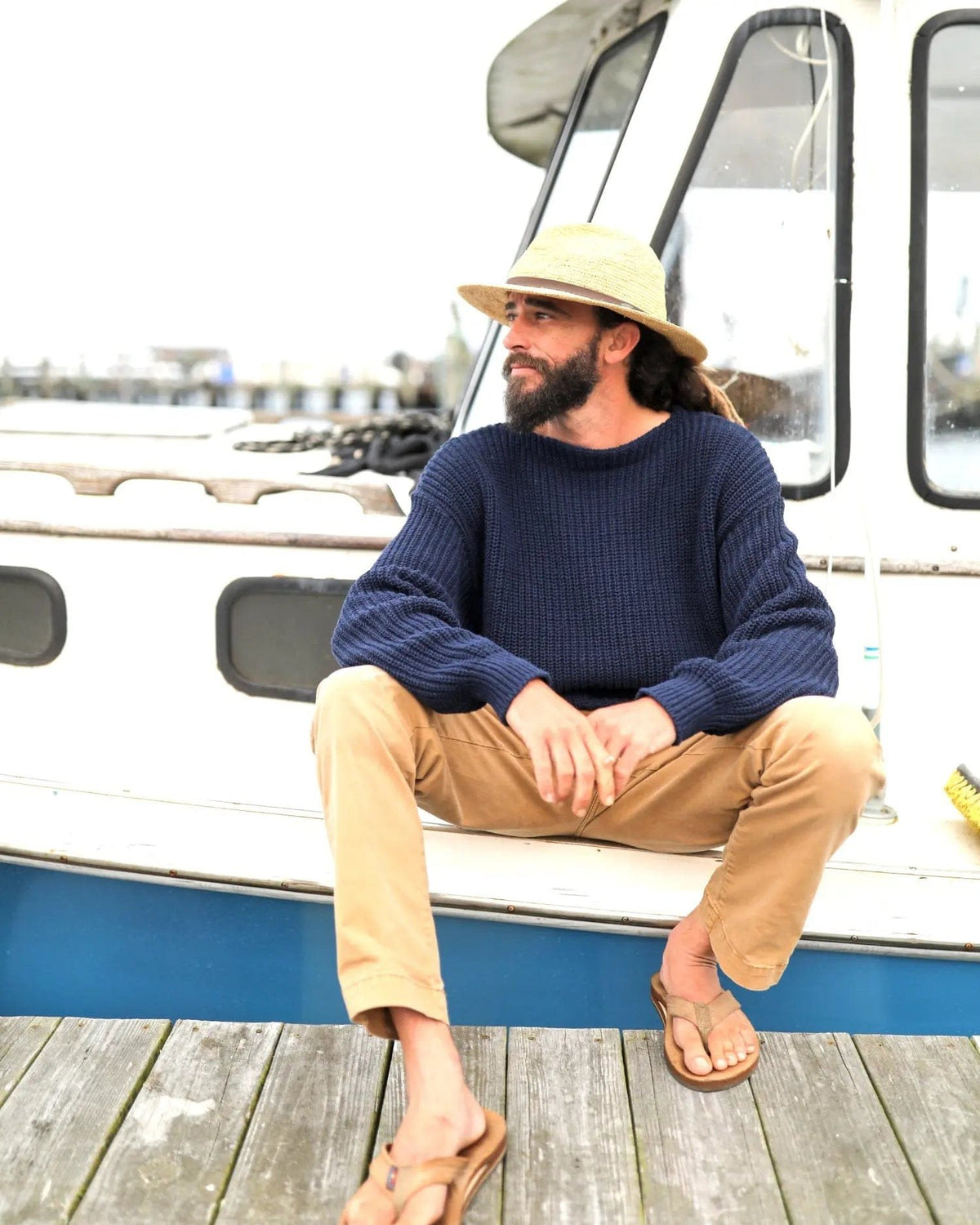 Whalerknits Newport Shaker Boatneck Sweater: A man in navy, tan trousers and a straw hat sits beside a boat cabin.