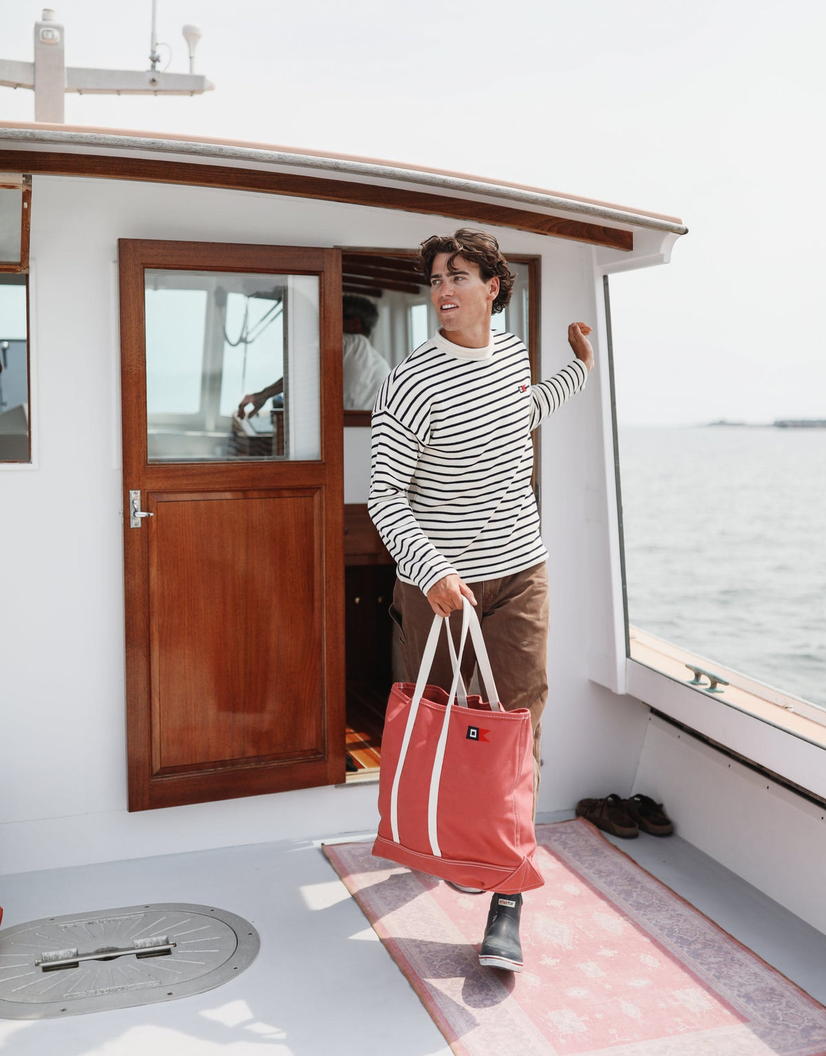 A man in a Signal Rollneck Sweater carries a Signal Medium Tote out of the boat's wooden cabin