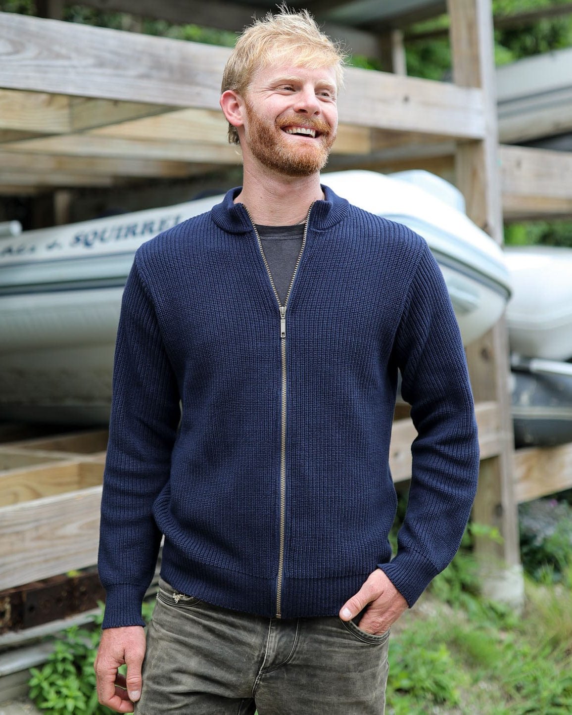 Whalerknits Cuttyhunk Full-Zip Cardigan: A man wears the navy cardigan with dark trousers beside stored boats.
