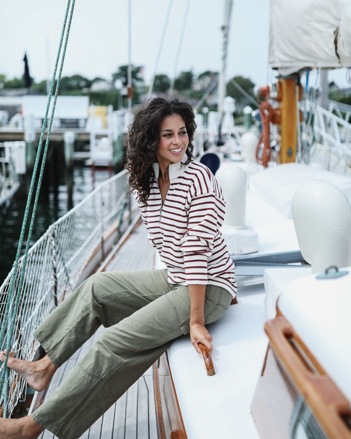 Whalerknits Nantucket Classic 1/4 Zip Sweater: A woman in natural with red stripes and olive trousers sits barefoot aboard a sailboat.