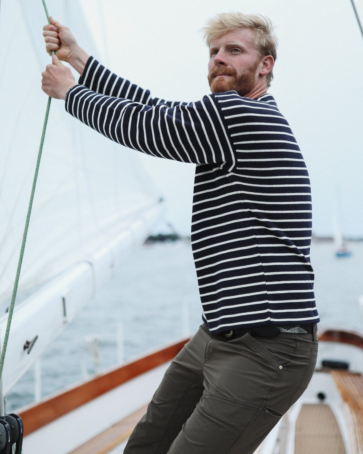 Whalerknits Nantucket Classic Boatneck Sweater: A man in navy with natural stripes pulls a rope aboard a sailboat.