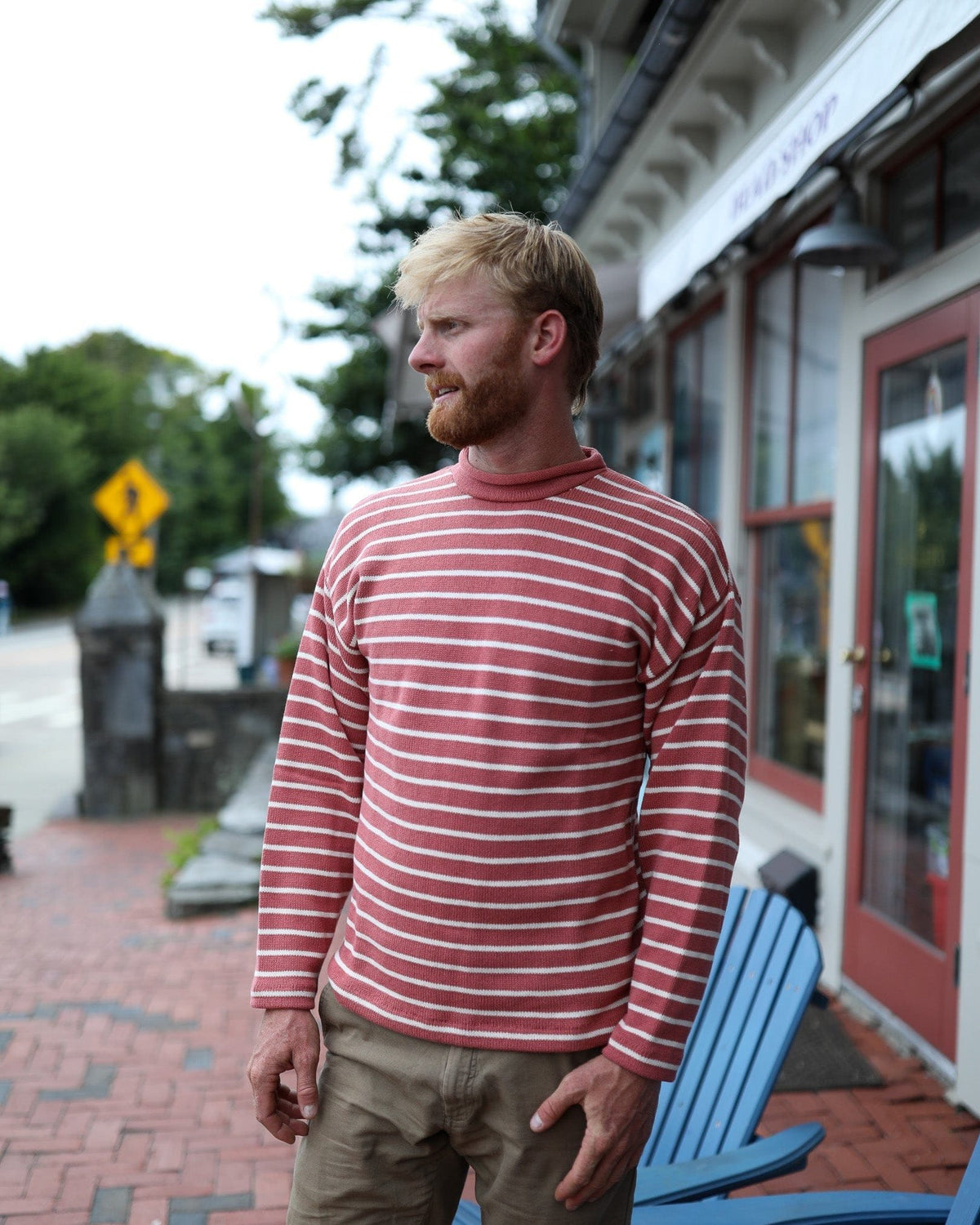 Whalerknits Nantucket Rollneck Sweater: A man in red with natural stripes stands on a brick sidewalk beside a shop.