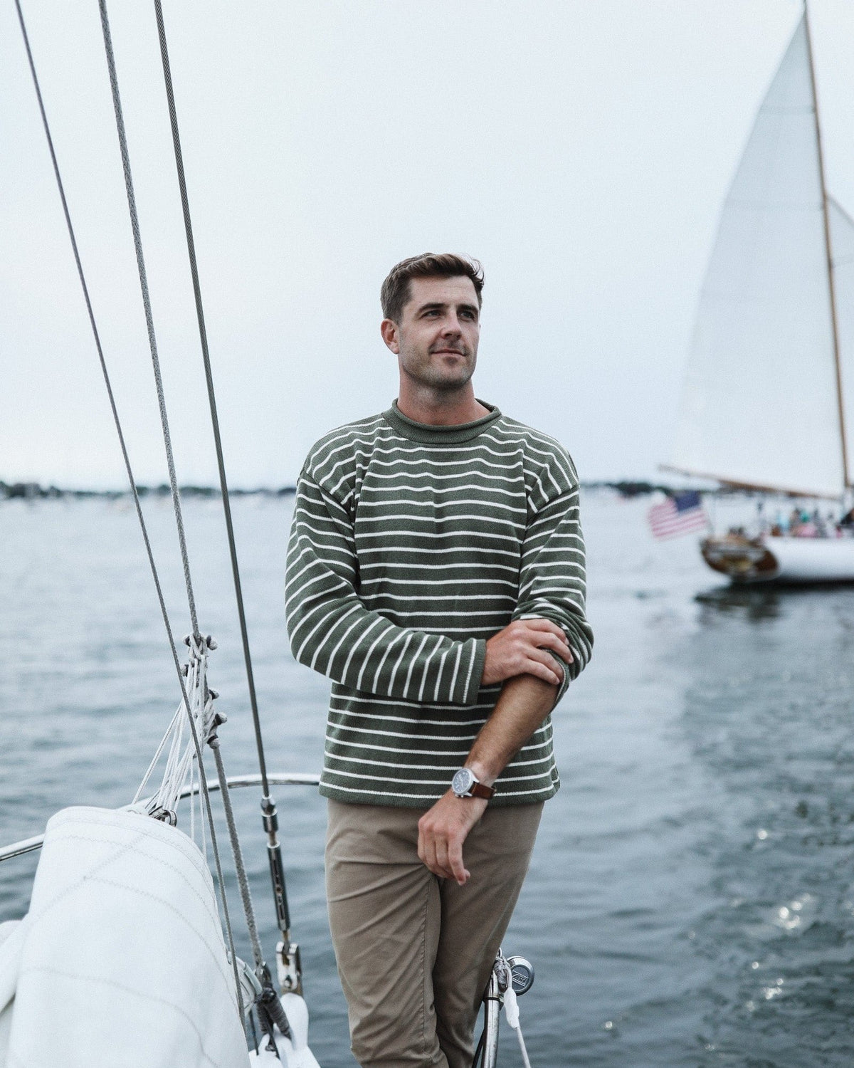 Whalerknits Nantucket Rollneck Sweater: A man in olive with natural stripes adjusts his sleeve aboard a sailboat.