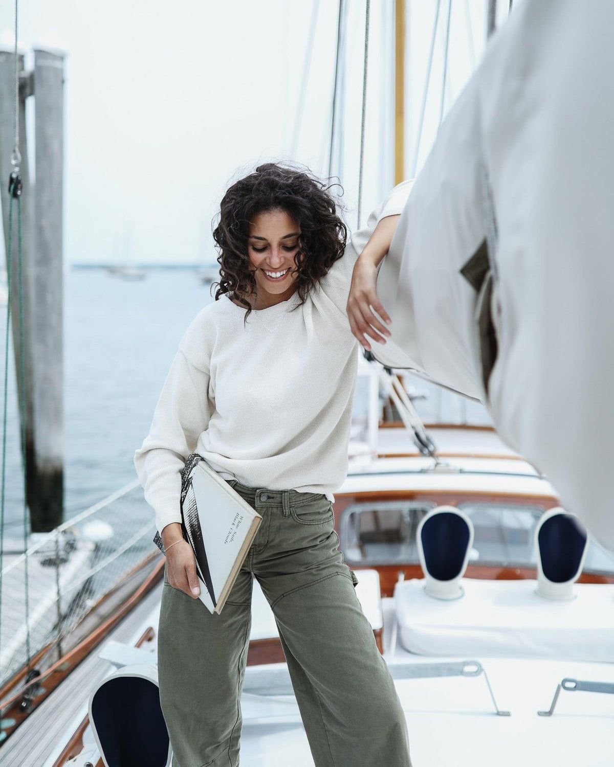 Whalerknits Chatham Jersey Sweatshirt Pullover: A woman in a light natural-colored pullover and olive trousers stands aboard a sailboat holding a book.