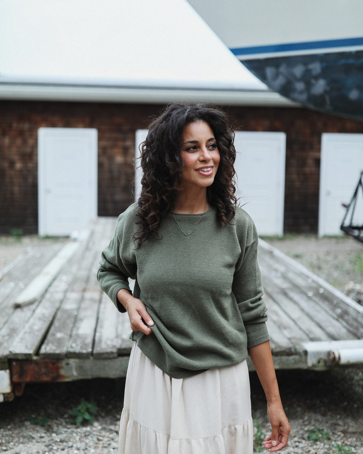 Whalerknits Chatham Jersey Sweatshirt Pullover: A woman wears olive with a light skirt beside a boatyard workbench. #Color_Olive