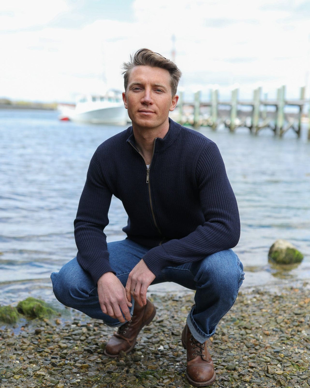 Whalerknits Cuttyhunk Full-Zip Cardigan: A man in navy and jeans crouches on a rocky harbor shoreline.