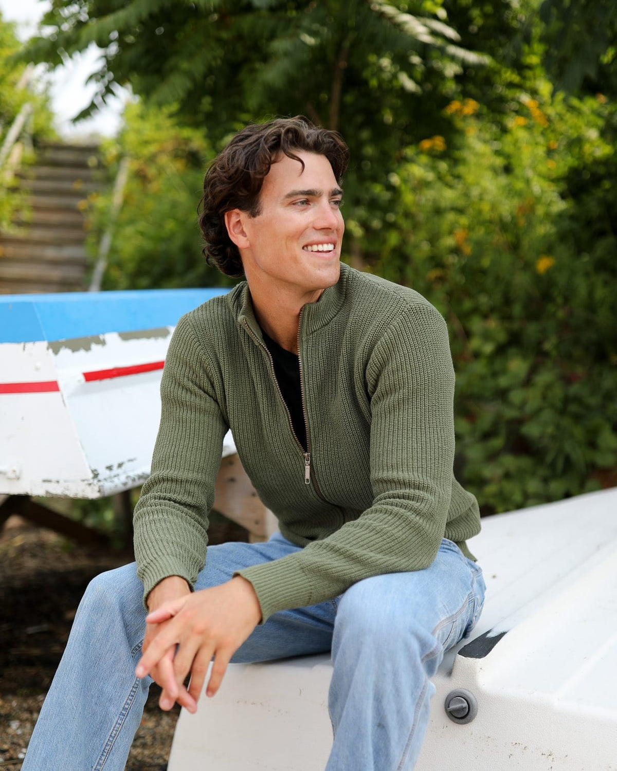 Whalerknits Cuttyhunk Full-Zip Cardigan: A man in olive and jeans sits on a small boat in a boatyard.