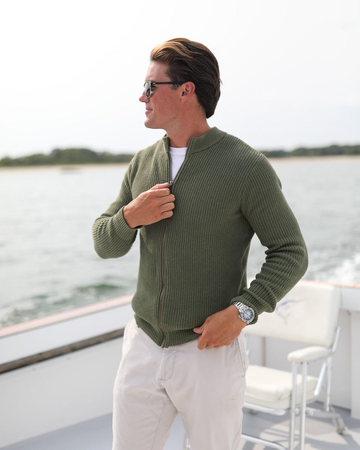 Whalerknits Cuttyhunk Full-Zip Cardigan: A man in olive and white trousers adjusts the zipper aboard a boat.