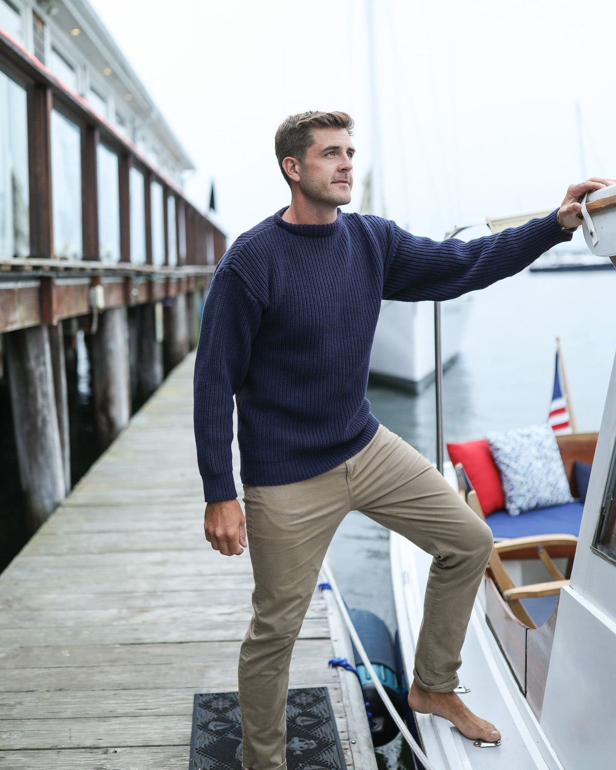 Whalerknits Newport Shaker Boatneck Sweater: A man in navy and tan trousers stands between a dock and sailboat, holding the rail.