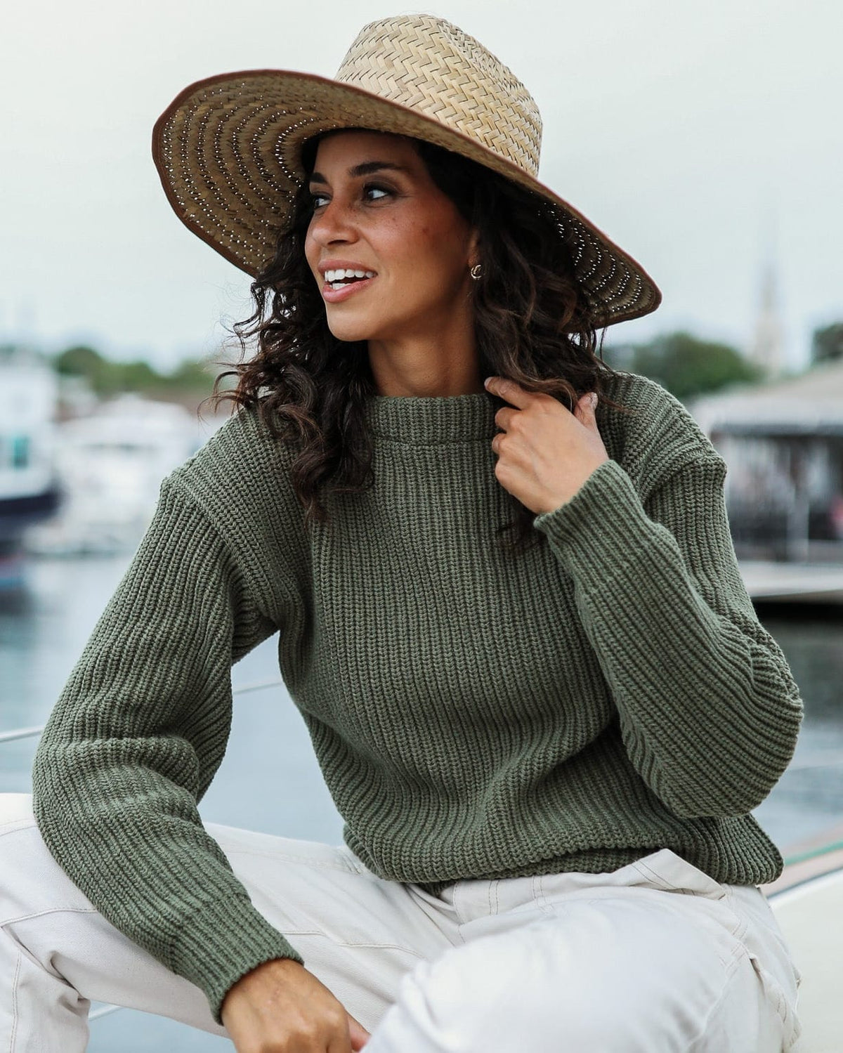 Whalerknits Newport Shaker Boatneck Sweater: A woman in olive, white trousers and a straw hat sits aboard a sailboat.