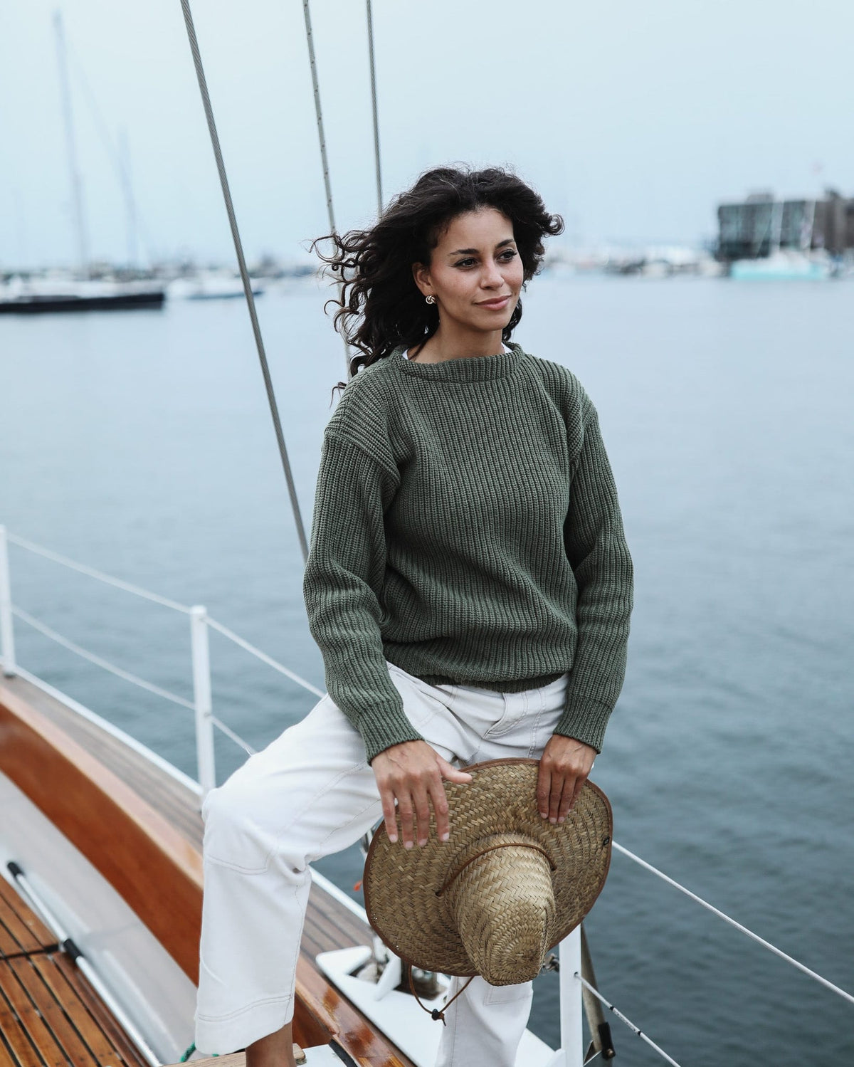 Whalerknits Newport Shaker Boatneck Sweater: A woman in olive and white trousers holds a straw hat while standing aboard a sailboat.