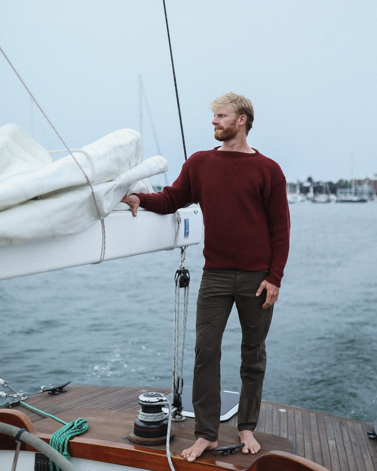 Whalerknits Newport Shaker Boatneck Sweater: A barefoot man in burgundy holds the covered boom aboard a sailboat.