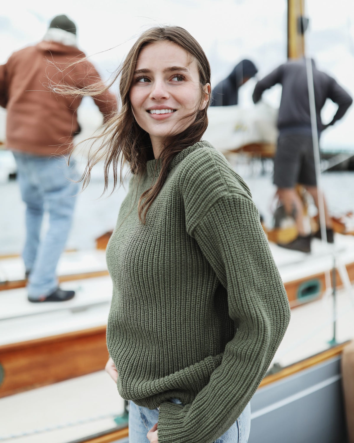 Whalerknits Newport Shaker Boatneck Sweater: Side view of a woman in olive and jeans smiling beside a sailboat.