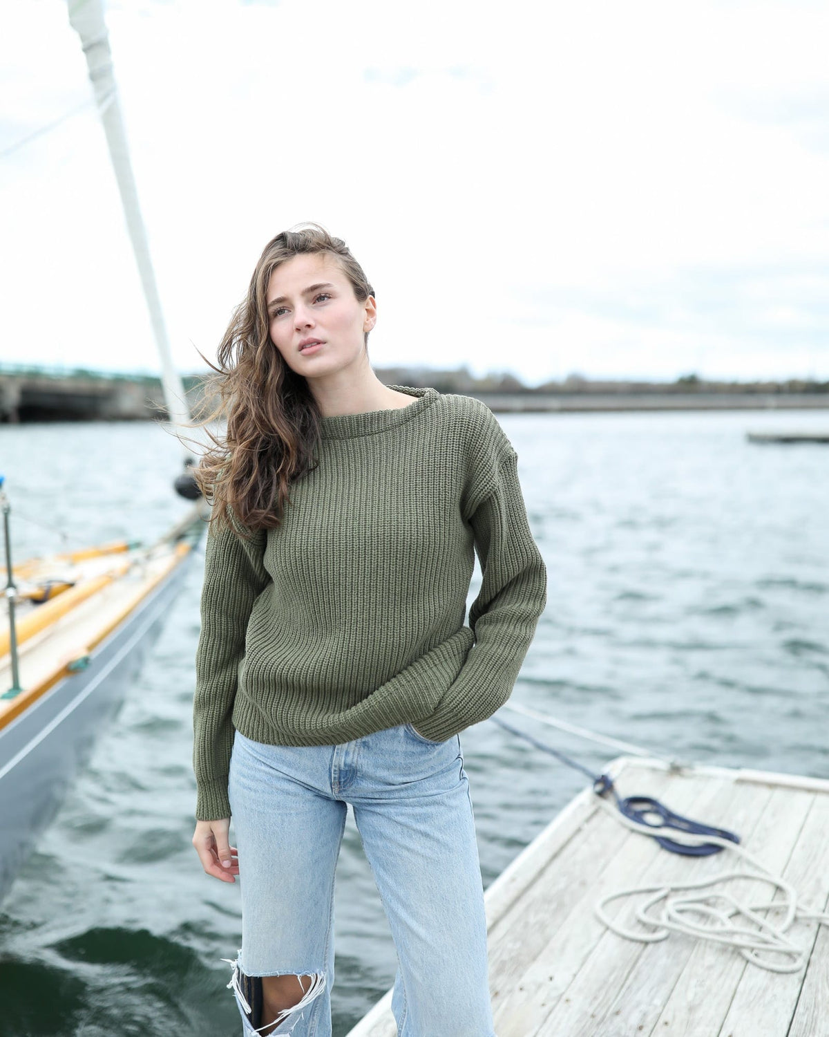 Whalerknits Newport Shaker Boatneck Sweater: A woman in olive and jeans stands on a dock beside a sailboat.