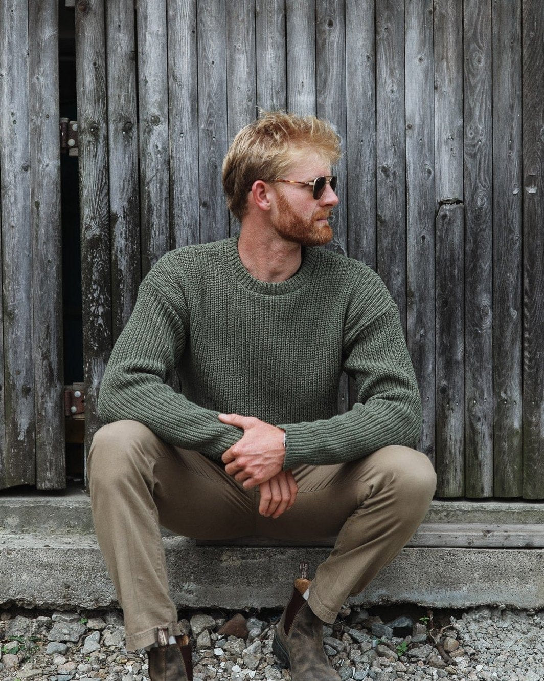 Whalerknits Newport Shaker Knit Crewneck Sweater: A man in olive and tan trousers sits against weathered wooden siding.