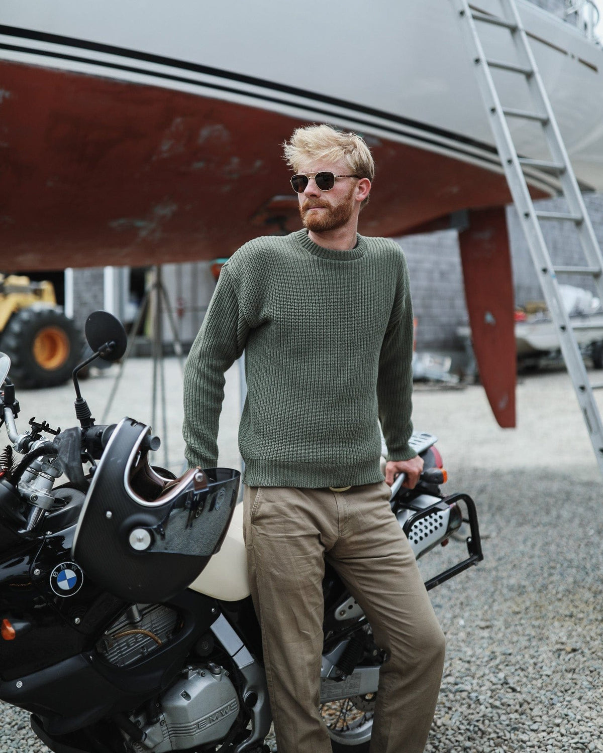 Whalerknits Newport Shaker Knit Crewneck Sweater: A man wears olive with tan trousers beside an outboard motor in a boatyard.