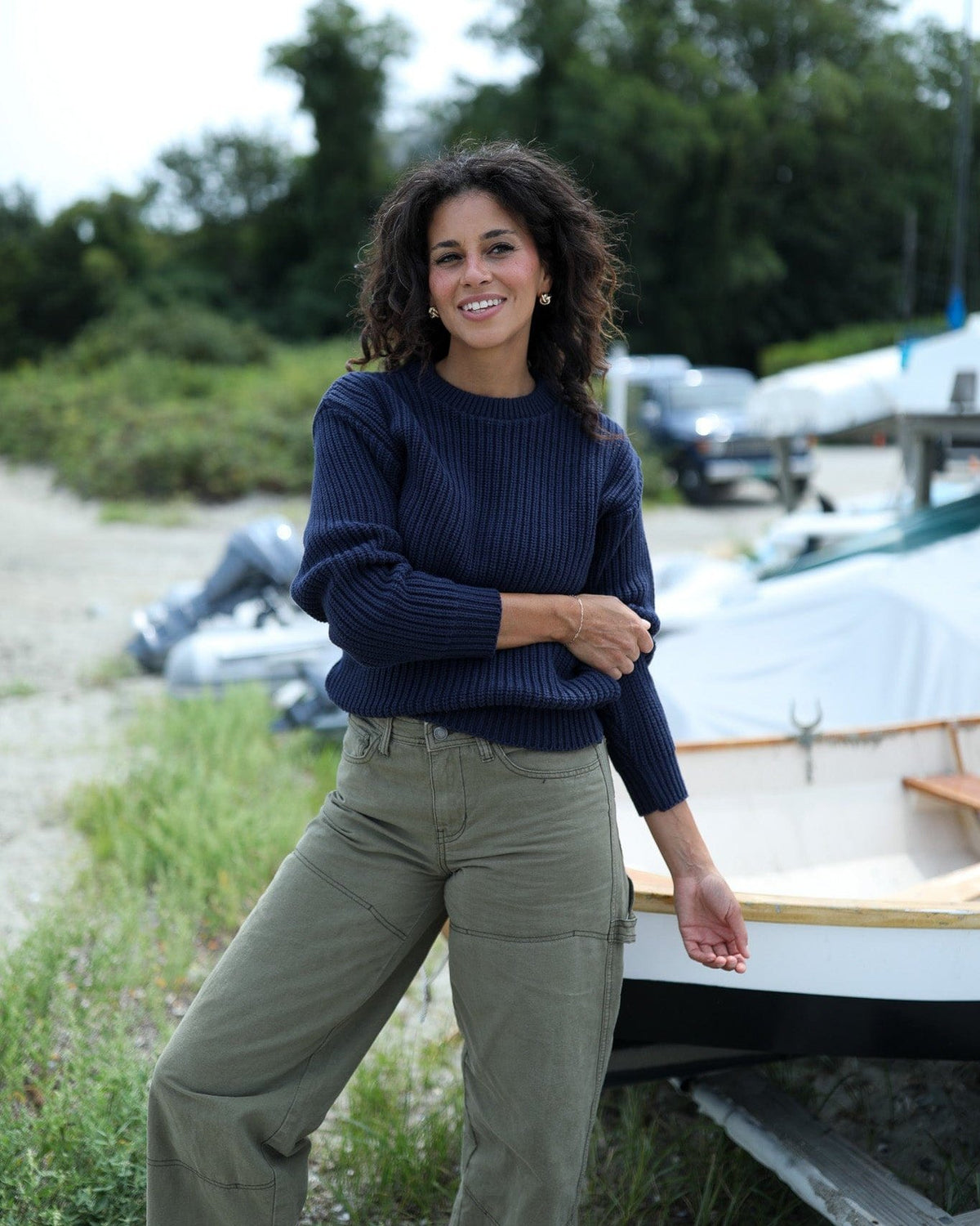 Whalerknits Newport Shaker Knit Crewneck Sweater: A woman in navy with olive trousers stands beside a small boat.