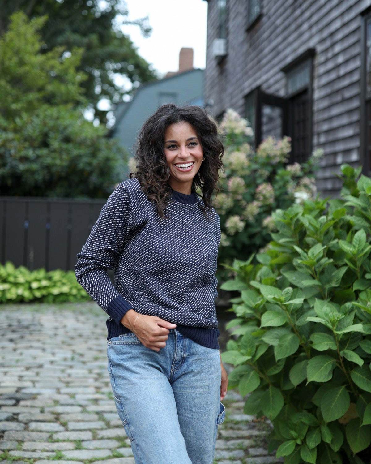 Whalerknits Sakonnet Birdseye Crewneck Pullover: A woman in navy with natural birdseye pattern and jeans stands beside a flowering shrub.