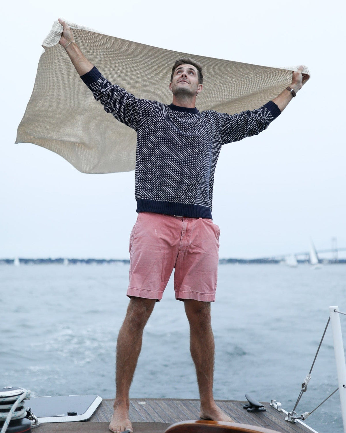 Whalerknits Sakonnet Birdseye Crewneck Pullover: A man in navy with natural birdseye pattern holds fabric overhead aboard a sailboat.