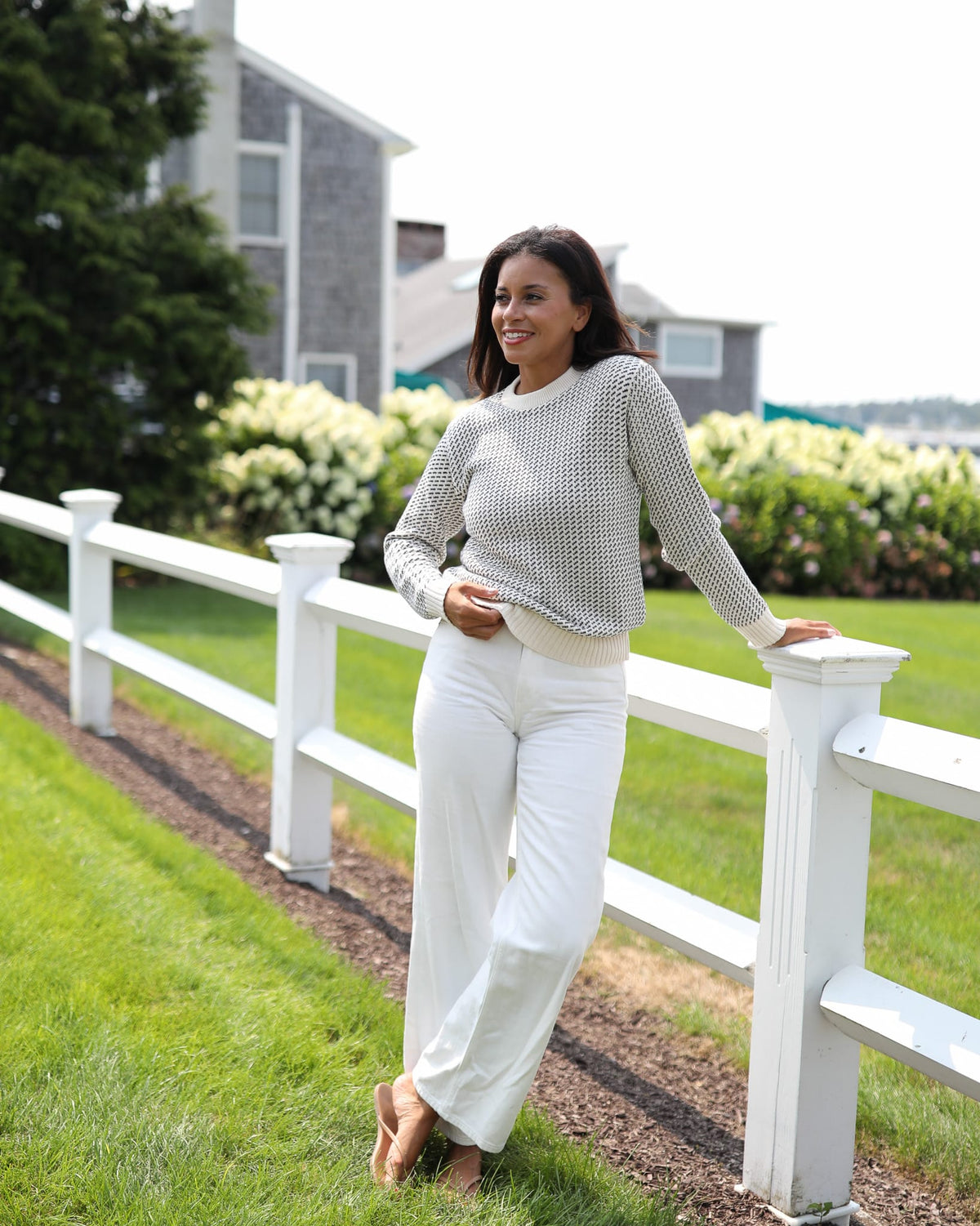 Whalerknits Sakonnet Birdseye Crewneck Pullover: A woman in natural with navy birdseye pattern and white trousers leans against a fence.