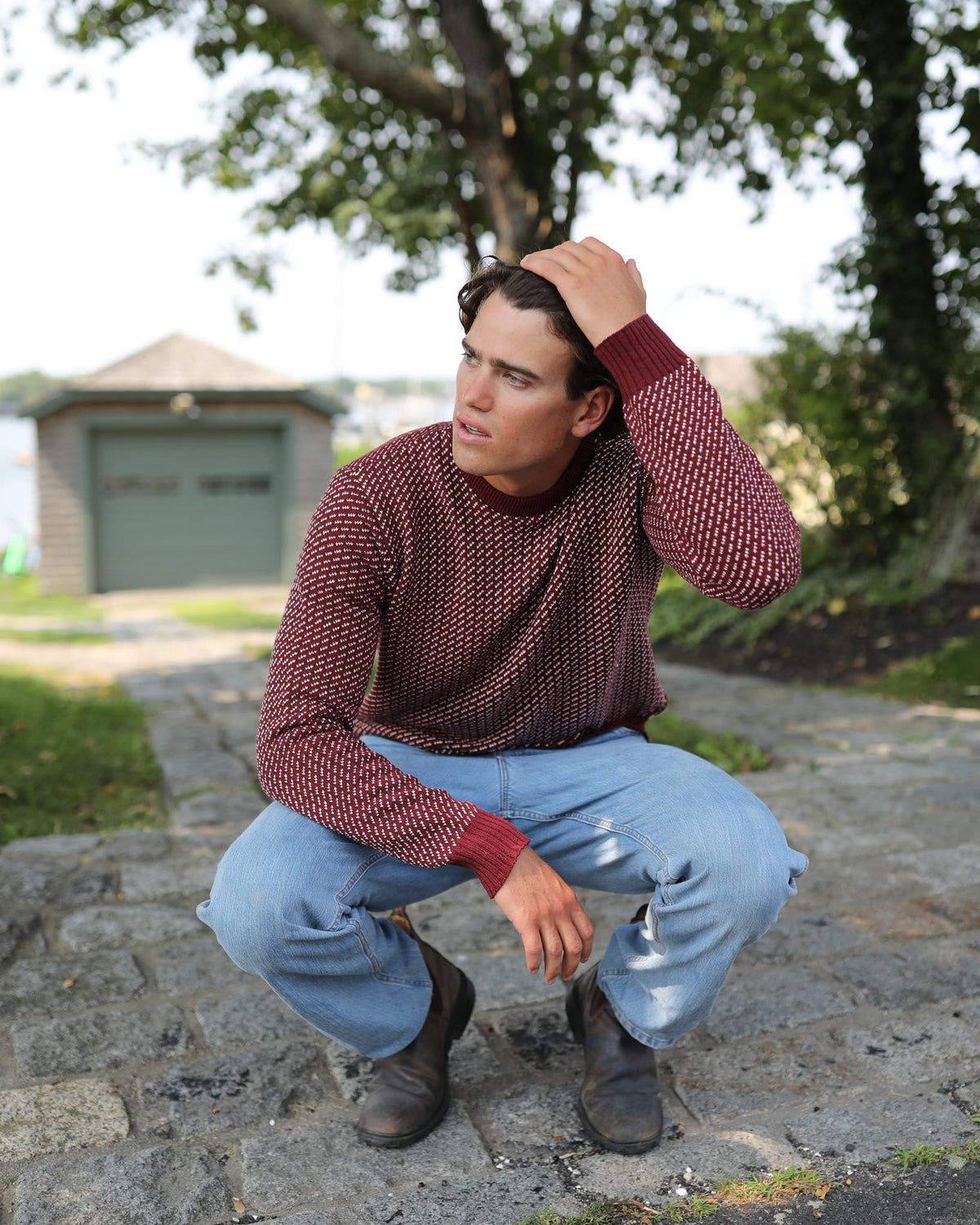 Whalerknits Sakonnet Birdseye Crewneck Pullover: A man in burgundy with natural birdseye pattern and jeans sits on stone steps with a hand in his hair.